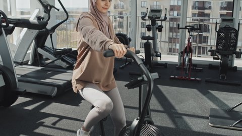 Tilt down shot of Muslim woman in hijab and sportswear using stationary bike while having cycle workout at gym