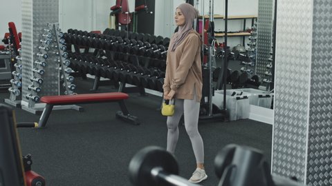 Muslim woman in hijab and sportswear doing weighted squats with kettlebell during gym workout