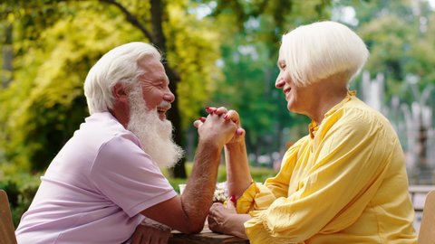 Pretty old woman and her husband smiling large at the cafe in the middle of park holding hands and looking with passion at each other