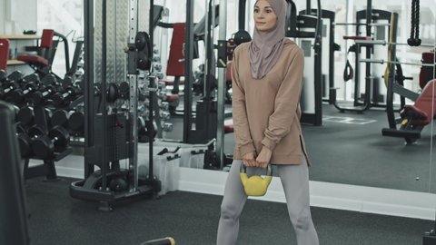 Muslim woman in hijab and sportswear holding kettlebell and doing squats while training at gym