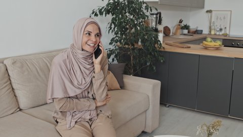 Zoom in shot of cheerful Muslim woman in hijab sitting on sofa at home, smiling and talking on mobile phone