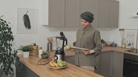 Muslim woman in hijab reading recipe on digital tablet and taking green apple while standing in kitchen at home