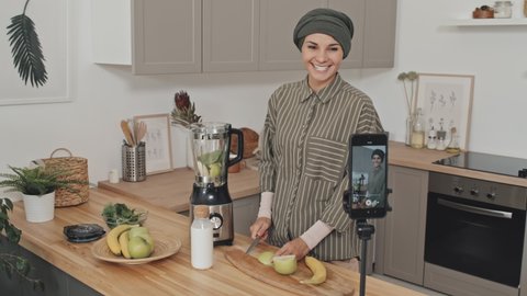 Muslim female blogger in hijab cutting fruit and talking on camera while filming video recipe with smartphone in kitchen