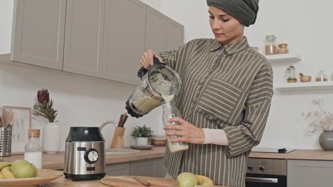 Muslim woman in hijab pouring fresh smoothie from blender into bottle in kitchen at home