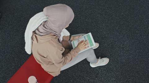 Top down shot of Muslim woman in hijab sitting in gym, wiping face with towel and checking physical activity statistics in fitness app on digital tablet