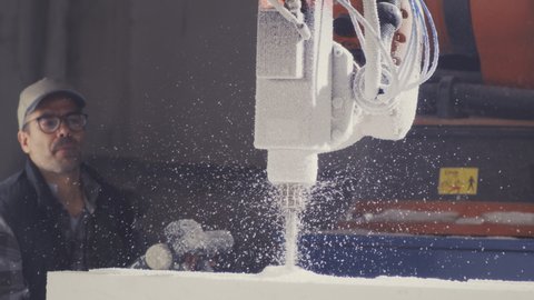 Craftsman in cap and glasses using remote control system and blowing off dust from polymer block with sprayer while cutting polystyrene with robotic arm milling machine in workshop