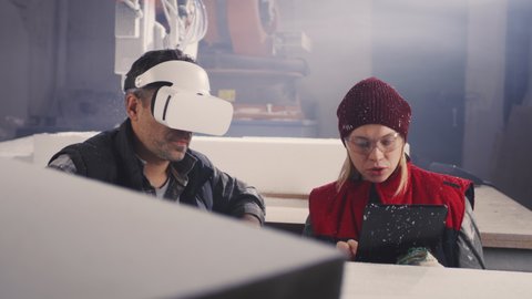 Middle aged man lifting VR headset and pointing at plastic block while female colleague entering data into tablet during work in modern workshop with robotic arm milling machine