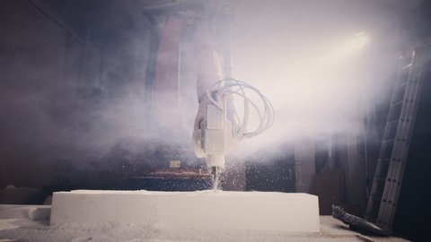Time lapse of robotic arm milling machine cutting block of polystyrene in cloud of smoke near craftsman in respirator and face shield