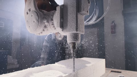 Pan around view of male employee in face shield and respirator watching robotic arm milling machine cutting polymer block during workday in daytime