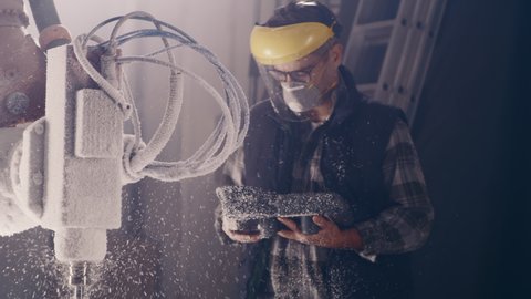 Craftsman in respirator and protective face shield using remote control system and watching milling machine cutting block of polystyrene in workshop