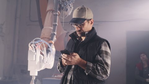 Mature craftsman in cap and glasses sending text message on cellphone while standing near colleague and robotic milling machine in modern workshop
