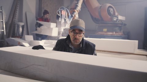 Middle aged male employee in cap and glasses examining and putting polymer block on workbench while female colleague cutting polystyrene with robotic arm milling machine in workshop