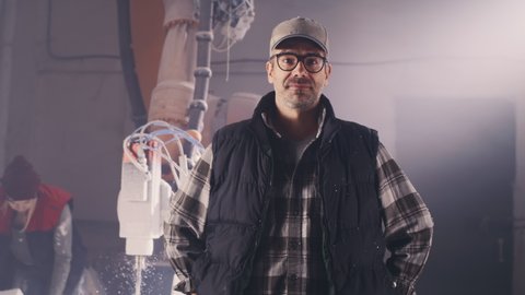 Self assured mature male artisan in casual outfit with glasses and cap holding hands on waist and looking at camera while standing near woman cutting polymer blocks with robotic arm milling machine