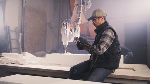 Middle aged male worker using remote control system and blowing off dust from plastic block while sitting on workbench near robotic arm milling machine