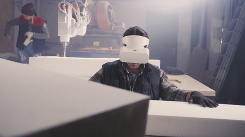 Male worker in modern VR glasses comparing plastic block with virtual plan while working in professional workshop with female colleague using robotic arm milling machine