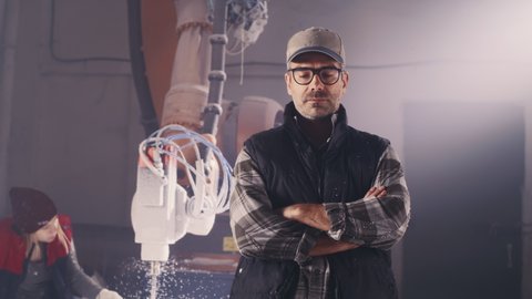 Pensive middle aged craftsman with crossed arms thinking over project while standing near female colleague working with robotic arm milling machine