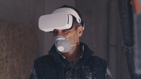Tracking shot of mature male artisan in respirator putting on VR helmet and examining virtual scheme near machine cutting polymer in workshop