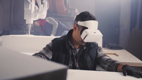 Mature man in contemporary VR headset touching and examining polymer block against colleague cutting polystyrene with robotic arm milling machine on workbench