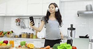 Low angle of cheerful pregnant female in headphones listening to music and dancing while shooting selfie video on smartphone in kitchen