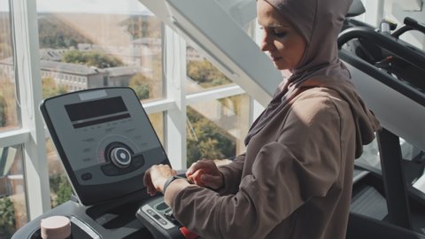 High angle shot of Muslim woman in hijab standing on treadmill in gym and using smartwatch