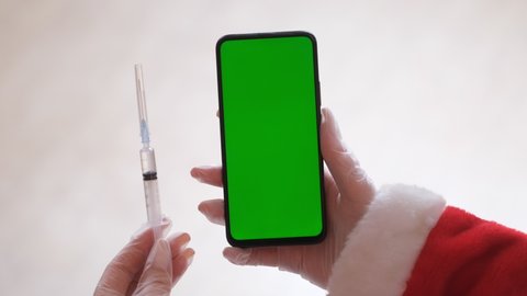 Vaccinated Santa Claus sitting in chair, showing mobile phone with green screen. Christmas spirit, holidays and celebrations concept 4k footage