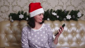 Emotional middle aged woman in pajamas and Santa hat having video call to friends sitting on the bed decorated Christmas branches
