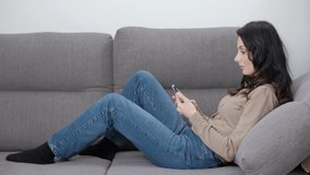Woman browsing internet on mobile phone while lying on comfortable couch in living room at home. 4K stock video clip of beautiful middle aged female model using modern smartphone to read news
