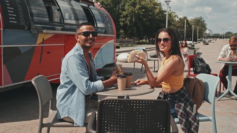 Medium portrait of young cheerful couple in sunglasses with tacos in hands sitting at table by truck with street food on summer day smiling at camera