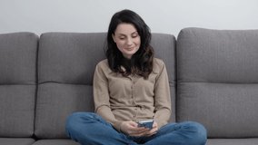 Happy young woman browsing mobile app on smart phone while sitting on couch at home. Stock video clip of attractive brunette model in 30s using social media application with cheerful smile 