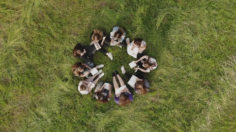 A group of 11 girls are sitting with notebooks on the grass in the park. Student work.