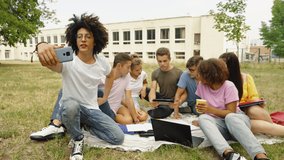 Black man with Afro hairstyle recording video with smartphone, his friends students sitting together on blanket, talking and using tablet and laptop, college building on background. Concept of blog