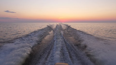 Sailing at calm Ladoga lake in sunset, thin stripe of sun seen at horizon, clear sky in beautiful shades. Camera look back from rushing vessel, waves from powerful motor split flat surface of water