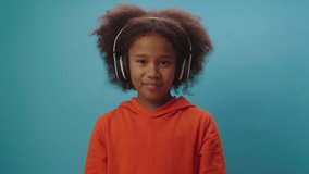 Confident African American girl in wireless headphones talking at camera standing on blue background. Child blogger speaking to camera recording video.