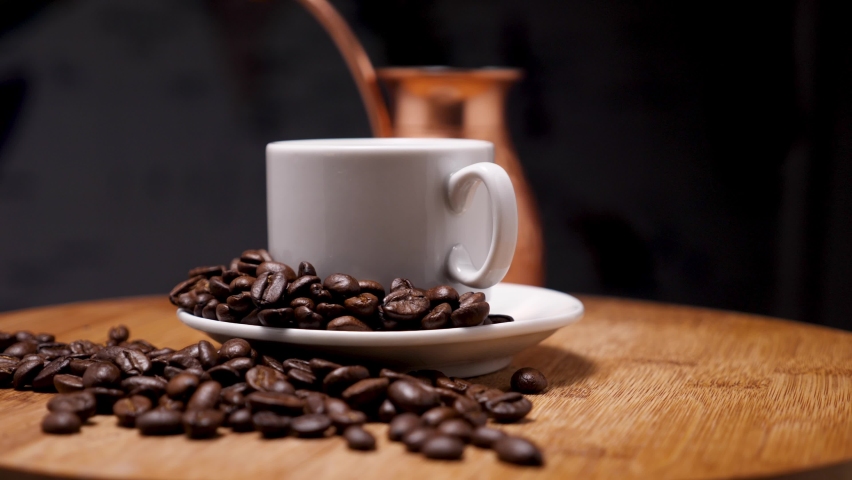Copper cezve and white coffee mug on a saucer on a wooden background. On the saucer and the board lie arabica coffee beans for making oriental coffee. The camera flies around. Parallax effect. Closeup | Shutterstock HD Video #1084038304