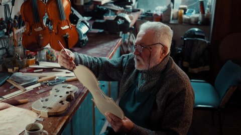 Old man shapes a wooden part for making a violin, a craftsman in his creative workshop works on making string instruments