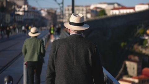 Elderly gray haired man in white fedora hat walks on Ponte de Dom Luis I. European tourism place in Portugal, city of Porto. Old traveling male tourist. Visiting Portuguese cityscape landmark sights.