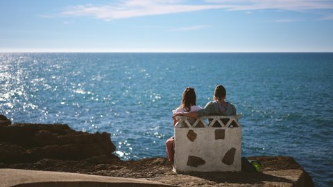 Tourism, scenery view at European Portuguese town of Ericeira. Young tourist people at sea. Travelers family sitting on stone bench by ocean. Lovely couple sea sights in Europe for traveling person.