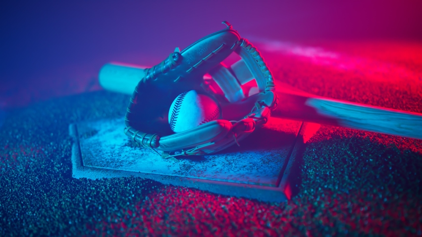 Baseball leather glove, ball and bat on a dirty white base in a red and blue lights. Dark stadium with orange gravel dirt and white base lines visible. National american sport equipment on the ground. Royalty-Free Stock Footage #1084041424