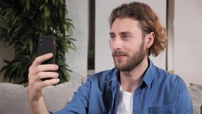 Positive young man making online video call chat interview with family using mobile phone app while sitting at home on sofa. Portrait of male blogger doing a videocall from his apartment.
