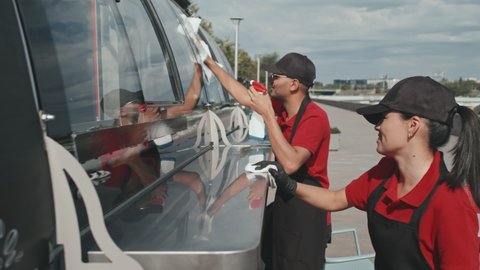 Medium shot of two young food truck workers in black caps and aprons cleaning its window and counter before opening on sunny day