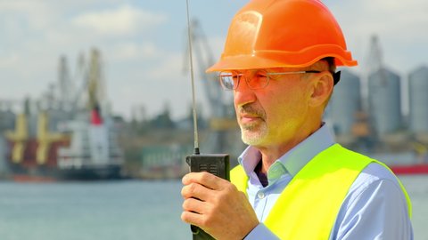 Sea industry port ship safety. Skilled inspector in orange hardhat commands loading process by portable radio in dockyard with tanks and cranes closeup