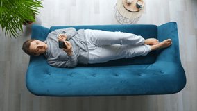 Overhead shot of happy relaxed woman holding smart phone, using mobile apps, watching funny video, having fun chatting in social media, lying on couch at home.