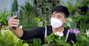 Portrait of a Happy young asian male gardener selling online on social media via smartphone in the garden. Man in face mask video call on mobile phone. Home greenery, selling online and hobby concept.