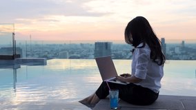 Asian Businesswoman with cocktail, sitting on edge at pool, reading email, typing response to client on laptop and woman hand closing lid of computer and enjoying view of Beautiful sunset in city.