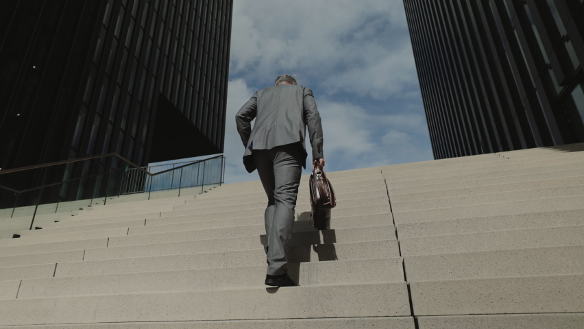 Low angle view of mature male entrepreneur in stylish formal clothes carrying suitcase while walking up on strairs. Way to work of businessman. Royalty-Free Stock Footage #1084066408