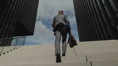 Low angle view of mature male entrepreneur in stylish formal clothes carrying suitcase while walking up on strairs. Way to work of businessman.
