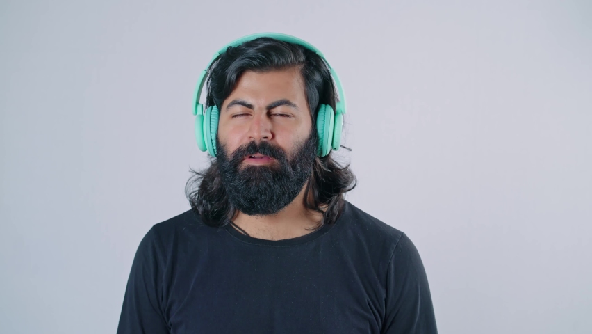 Modern young happy cheerful Indian Asian bearded male with dark black long hair wearing headphones dancing or grooving isolated on a white background screen. Concept of online music streaming services