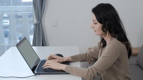 Freelance worker woman typing text on laptop keyboard while sitting behind white table in living room. Brunette model in 30s working from home with mobile computer