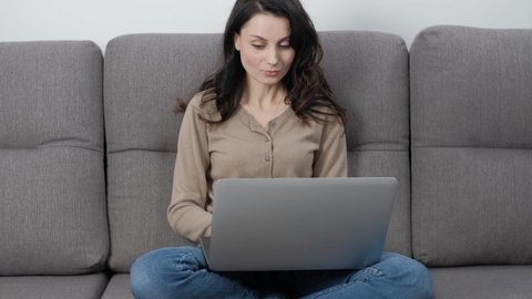 Stock video of freelancer woman working from home with laptop computer. Beautiful young adult model with long dark hair typing text on notebook keyboard while sitting on comfortable sofa in room 