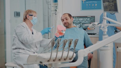 Dentist showing correct way to clean denture with toothbrush, holding artificial jaw for example. Stomatologist giving hygiene lesson to patient learning to brush teeth. Man preventing caries